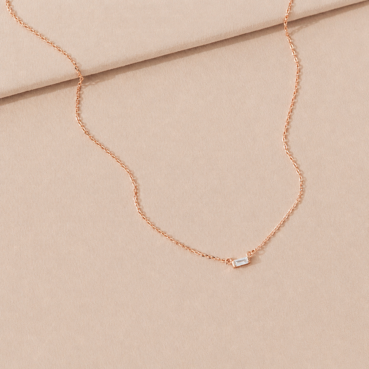 Rose gold necklace with a small rectangular pendant on a beige fabric background